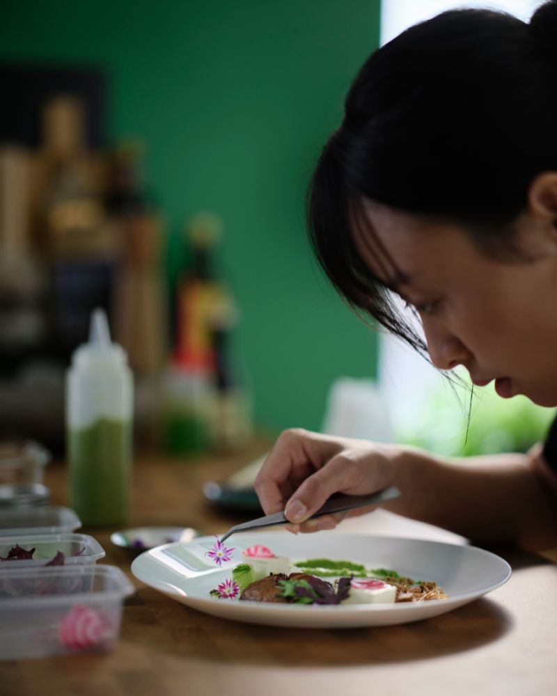 A person is leaning over a white OFTAST deep plate with food on it, carefully placing a fl ower on the food using tweezers. 