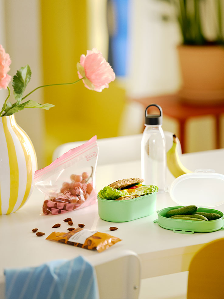 Una mesa con botella de agua IKEA 365+ en gris oscuro,  fiambrera apilable UTBJUDA en verde claro y almendras tostadas MUNSBIT.