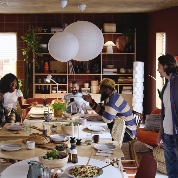 Light wood dining table in a living room with shelving. Hanging paper lamps. Social atmosphere with people eating.