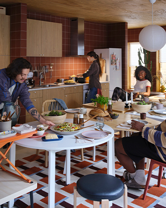 White extendable dining table. A man serves food. Kitchen with light wood cabinets and brown tiles.