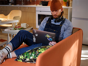 A man sitting in an orange velvet armchair, using a laptop. He is wearing a beanie and headphones.