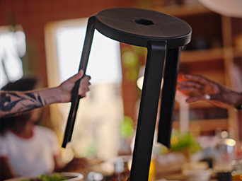 A dark wood stool with a hexagonal seat is held in the air. Blurred dining room atmosphere in the background.
