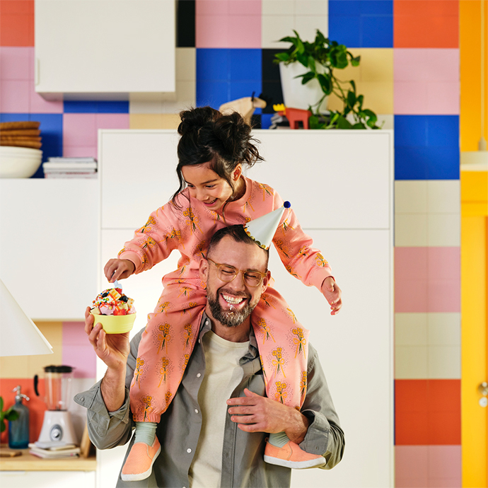 A smiling father carries his daughter on his shoulders in a colorful kitchen. The girl holds a bowl of fruit.