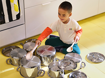 A boy sitting on the yellow floor of a kitchen, surrounded by metal pots and pans. He holds two wooden spoons.
