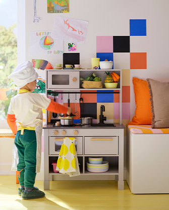 A boy dressed as a chef plays with a light gray wood toy kitchen. There are drawings on the wall and a bench with cushions.