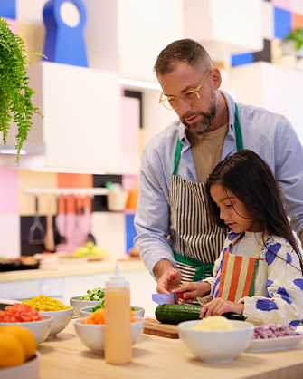 A father and daughter cut cucumber in a kitchen with multicolored tiles. Several bowls of ingredients on the wooden countertop.
