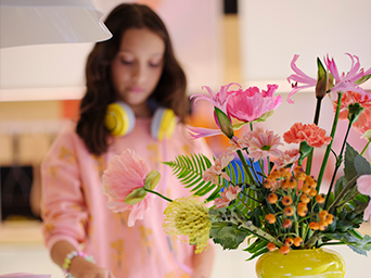 A bouquet of pink, orange, and yellow flowers in a vase. A girl with headphones and a pink sweatshirt appears blurred in the background.