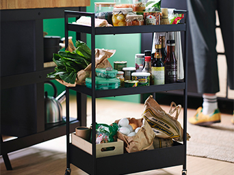 Three-tier black utility cart with food and products. Nearby, a kettle and a cabinet with white drawers.