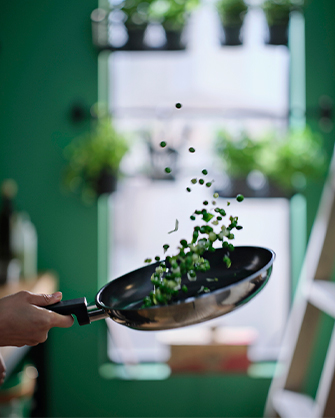 A person tosses peas in a black pan in the air. In the background, a kitchen with hanging pots and a ladder.