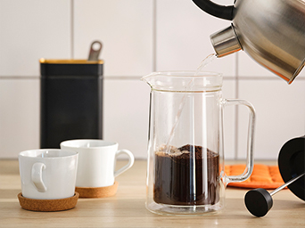 Primer plano de una prensa de café con jarra de cristal doble llenándose de agua caliente, con dos tazas y un recipiente al lado.