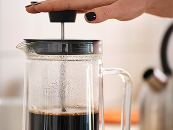 Primer plano de una prensa de café de cristal con el émbolo bajando para filtrar el café. Una mano con uñas negras empuja el émbolo.