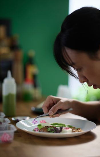 A person is leaning over a white OFTAST deep plate with food on it, carefully using tweezers to place a flower on the dish.