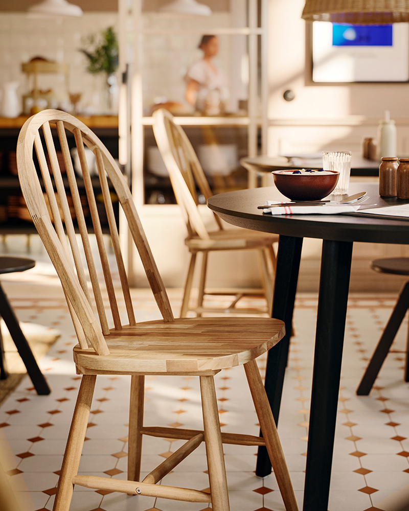 Una silla SKOGSTA de acacia maciza con respaldo arqueado y barrotes está junto a una mesa redonda oscura en un área de comedor comercial.