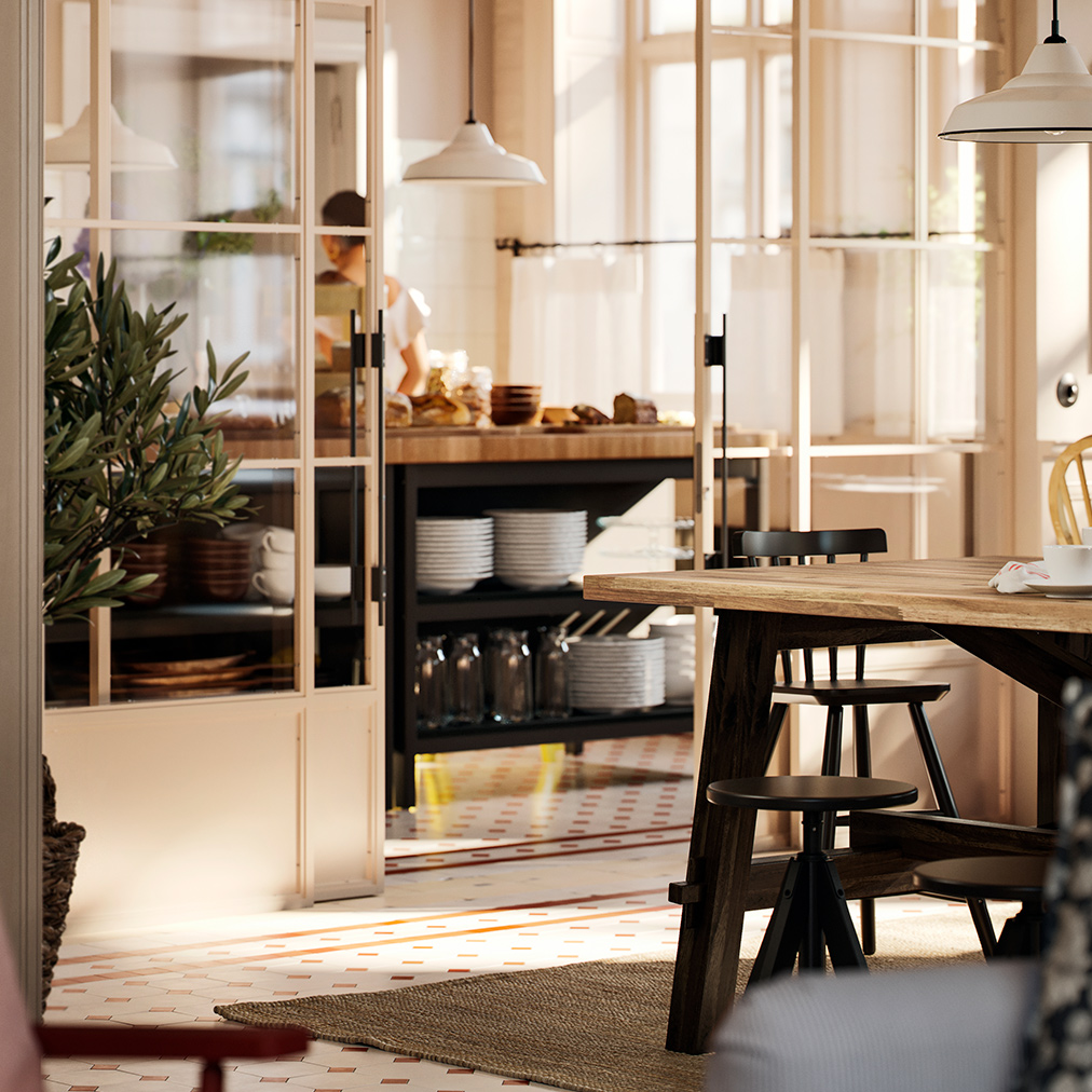 Una parte de una mesa de comedor SKOGSTA de acacia con patas negras es visible junto a un área de buffet de desayuno que está detrás de una pared de vidrio.