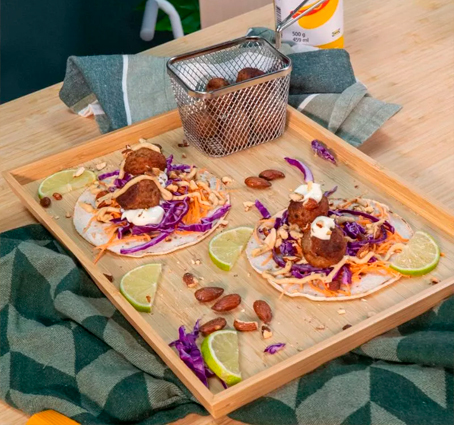 Tacos de albóndigas servidos en un plato de madera, cubiertos con col morada picada, zanahoria rallada, salsa blanca, almendras tostadas y perejil, acompañados de gajos de lima y una cesta metálica con más albóndigas.