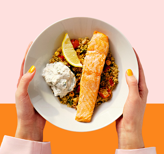 Plato de salmón y ensalada, sostenido por dos manos sobre un fondo rosa y naranja.