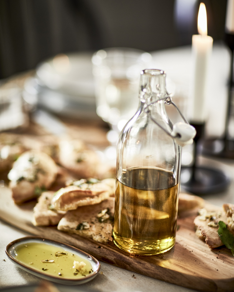 Botella de vidrio con aceite de oliva sobre tabla de madera, pan, velas y vajilla minimalista – mesa acogedora 