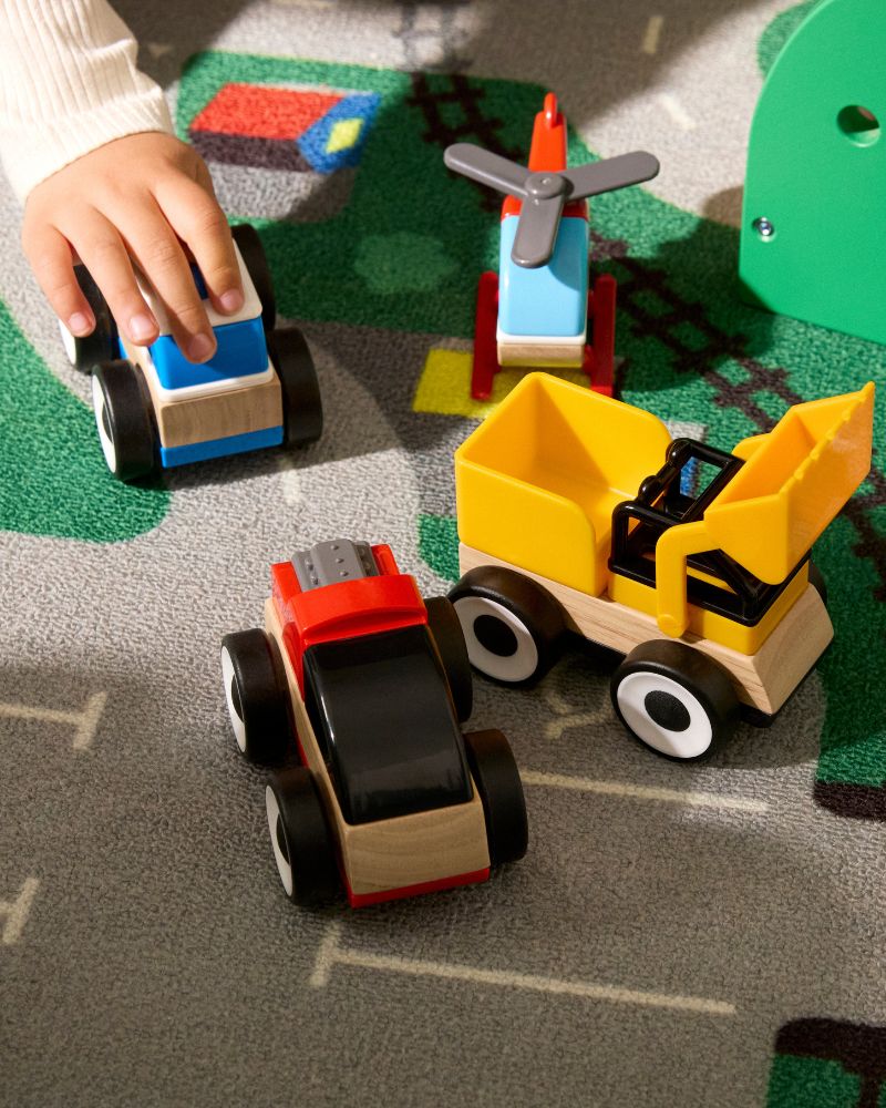 La mano de un niño juega con coches de juguete: un camión rojo y negro, una excavadora amarilla y un helicóptero azul.