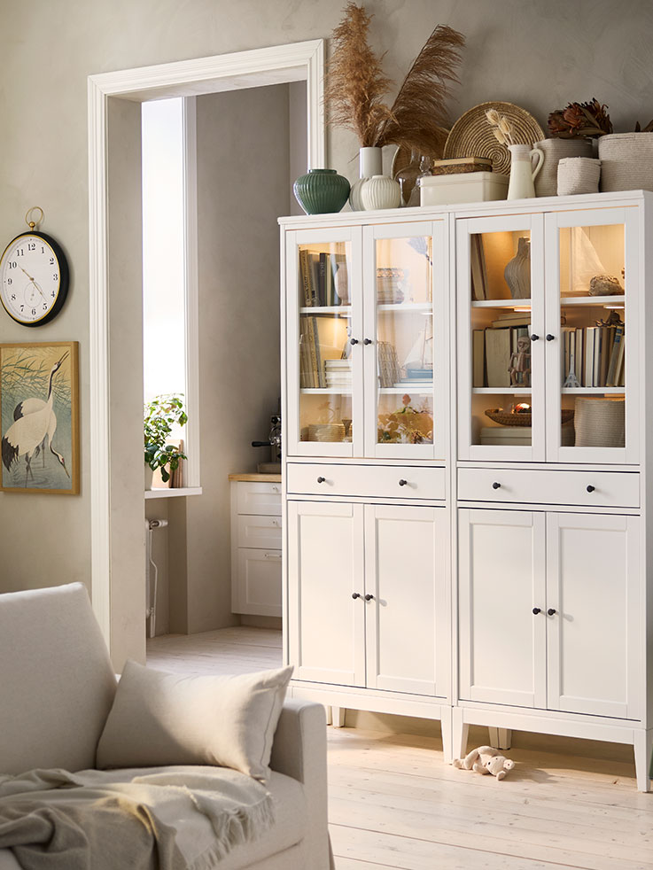 Two white IDANÄS high cabinets with glass doors and 1 drawer, placed side by side, have books and keepsakes stored inside