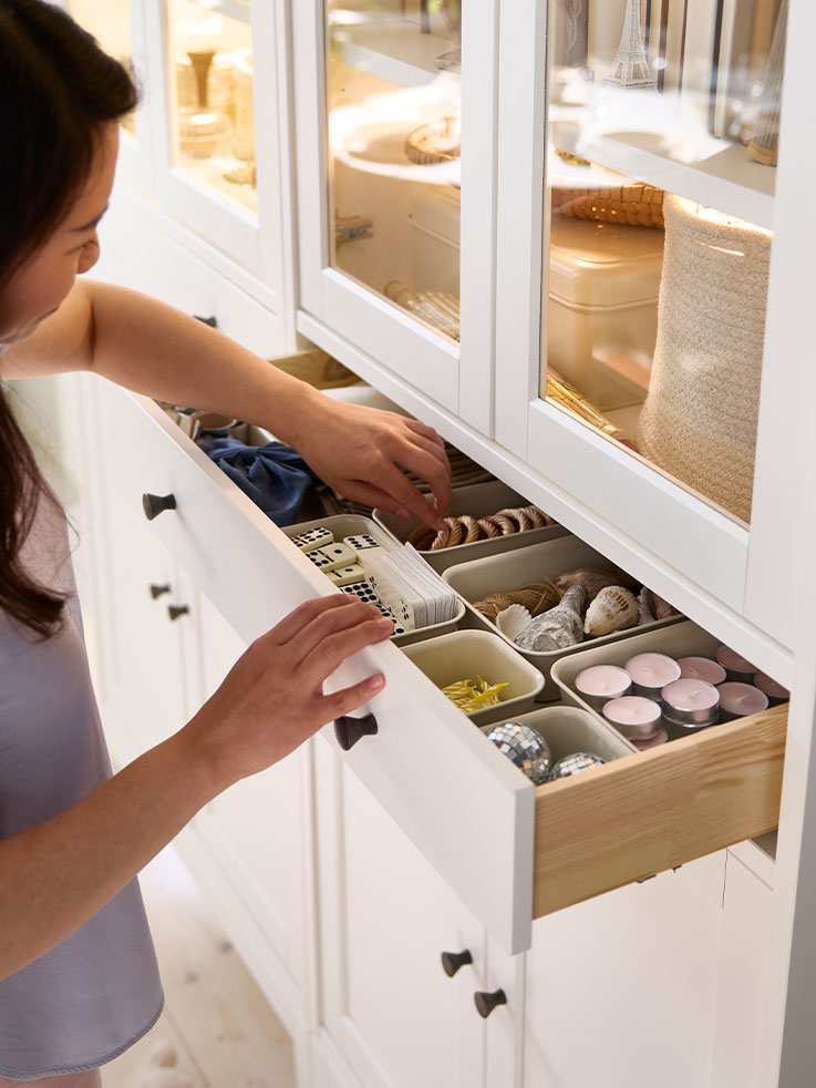 A person has pulled out the drawer in an IDANÄS high cabinet with glass doors and 1 drawer, reaching for items stored inside