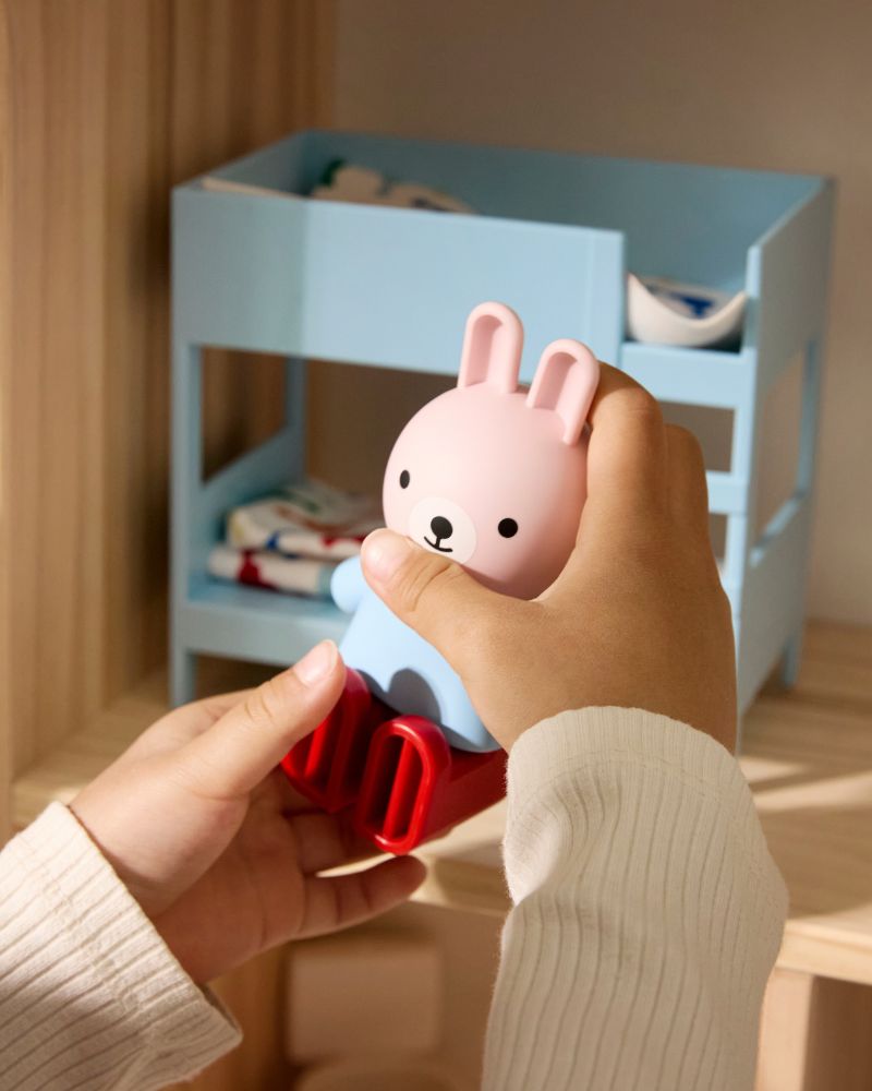 Las manos de un niño sostienen un juguete de conejo rosa y azul. Al fondo, hay una litera de casa de muñecas.