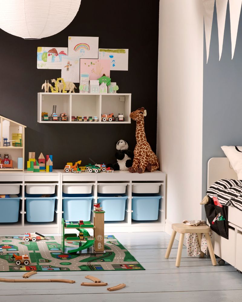 Un colorido cuarto de juegos infantil con una pared negra, muebles de juguete blancos con cubetas azules, y una jirafa.