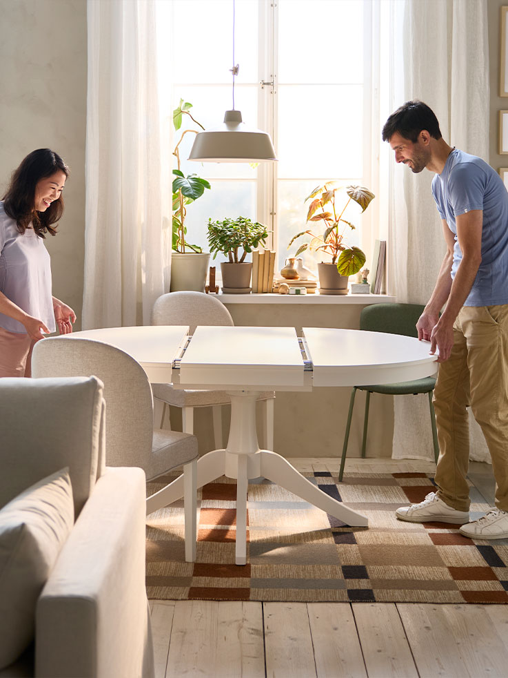 Two people are pulling out a round white ROSENTORP extendable table to make it bigger with the integrated butterfly leaf.