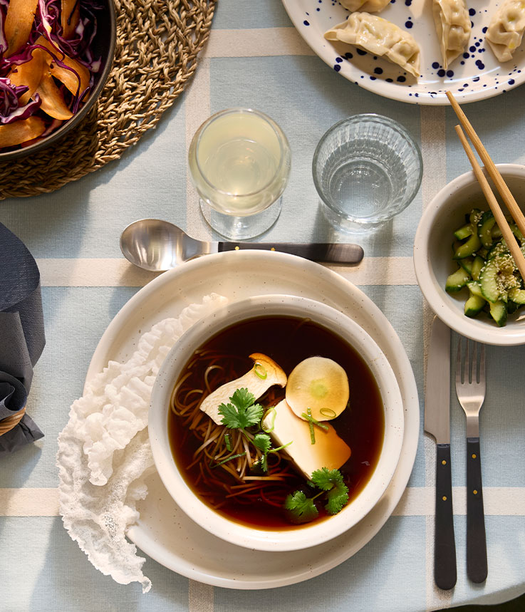 A dotted/light beige DAGGASTER plate and bowl has a soup made of mushrooms, tofu and noodles, next to a bowl of avocados.