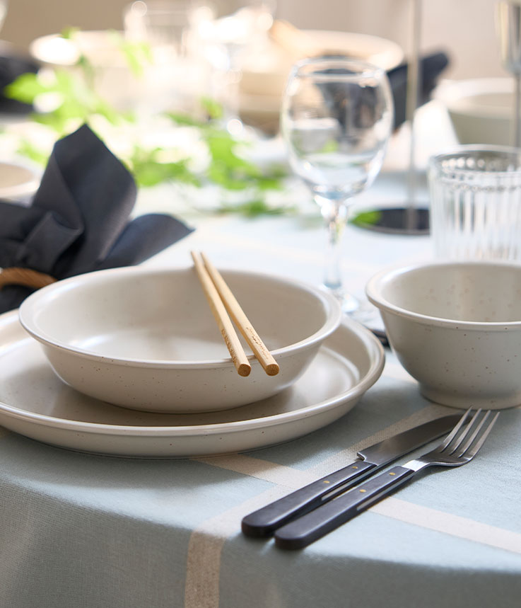 A dotted/light beige DAGGASTER bowl is placed on a plate, with two chopsticks on top of the bowl and cutlery on the side.