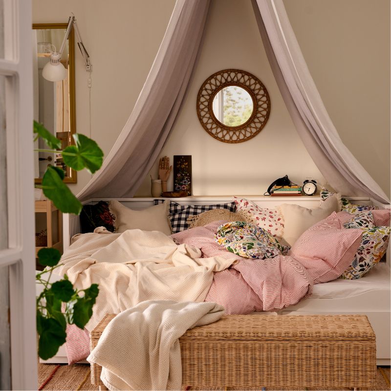 HEMNES day-bed with bedlinen in pink and white, TOLKNING rattan bench in foreground, a mirror and a textile canopy above bed.