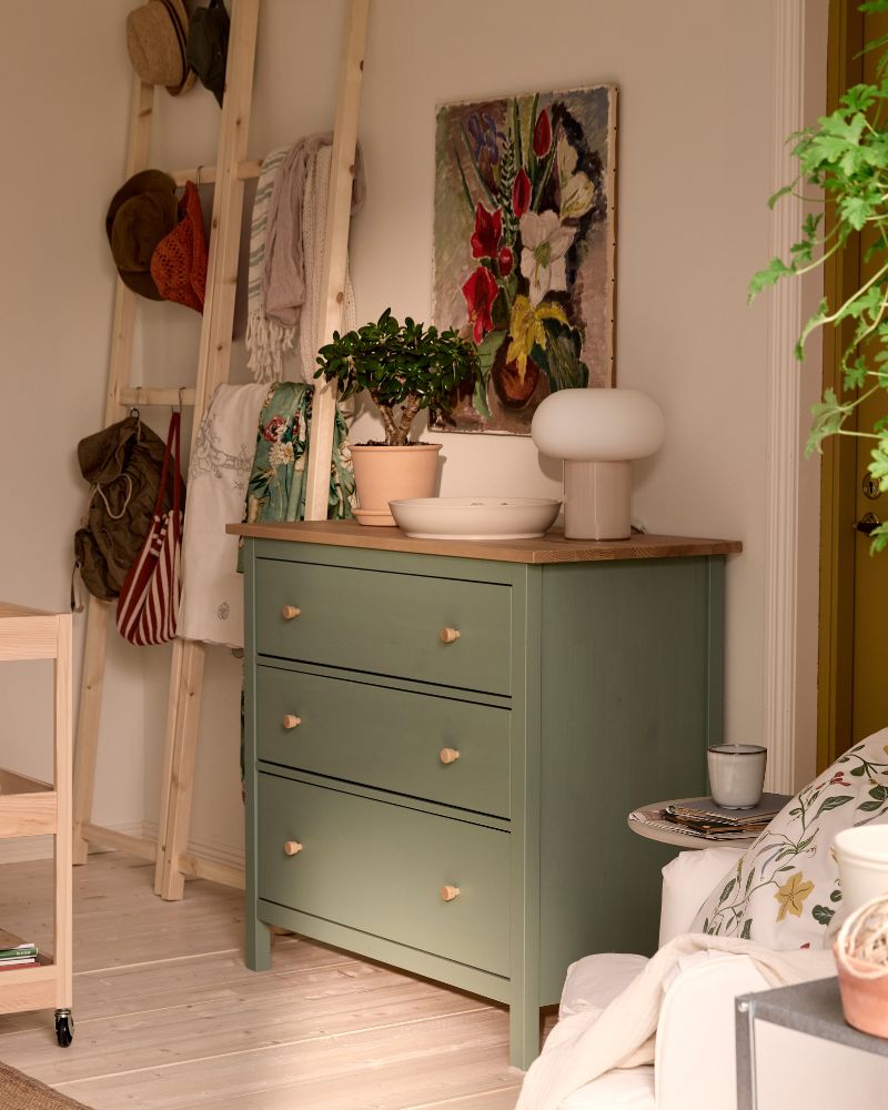 A green HEMNES chest of drawers with pots and plants on top, a picture above it and wooden ladders in the back for storage.