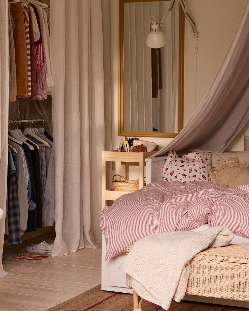 A room with a table to the left in the foreground and a bed to the right. A wardrobe solution with curtains in back.