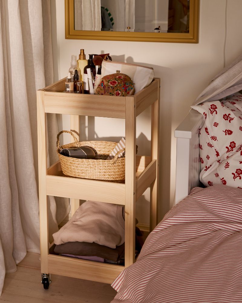 Close-up of LOSHULT trolley in pine next to a bed. A mirror above it, and the trolley is full of accessories and things.
