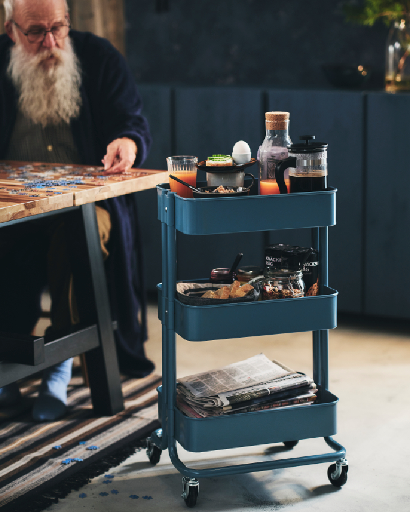 Un anciano con una larga barba coloca un rompecabezas sobre la mesa del comedor. A su lado hay un carrito RÅSKOG con su desayuno.