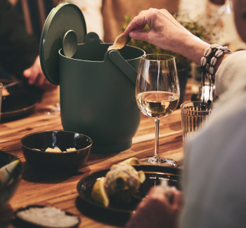Una anciano sentada en una cena coloca los restos de alcachofa en un elegante cubo de basura de mesa.