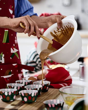 Una persona con delantal vierte la mezcla para pasteles en moldes festivos VINTERFINT, sobre una tabla de cortar, sobre una mesa.