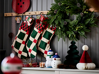 Calcetines navideños con diseño de cuadros verdes y rojos colgados de perchas de madera. Al lado, una corona de abeto con piñas.
