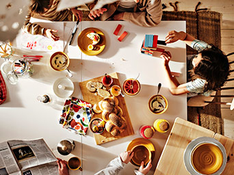 Vista cenital de desayuno compartido con pan, fruta y prensa. Varias personas alrededor de la mesa.