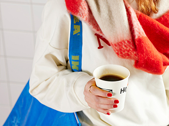 A person holds a takeaway coffee and carries a blue IKEA bag. The person is wearing a white sweater and a red checkered scarf.