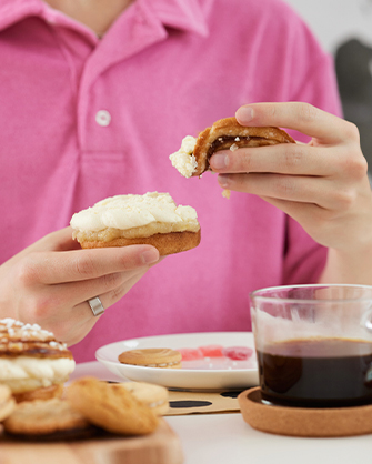 A person wearing a pink t-shirt holds two halves of a sweet bun with cream. There are cookies and sweets on the table.