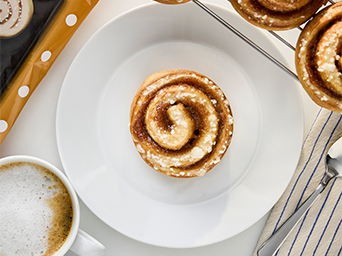 A cinnamon roll with frosting and sugar on a white plate on a table. To the side, there is a cup of latte.
