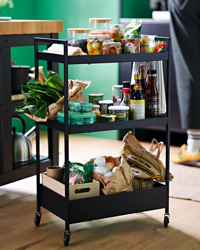 Carrito de cocina negro de tres niveles con ruedas. Almacena verduras, huevos, tarros de comida, y botellas de condimentos en una cocina.