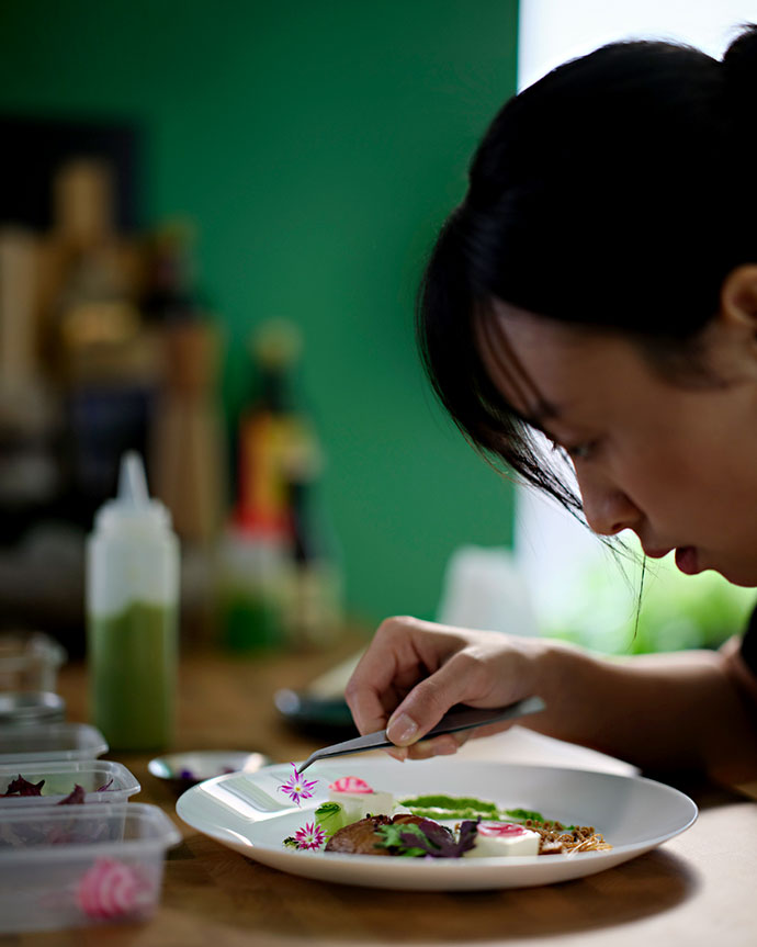 Una chef decora con precisión un plato blanco con flores comestibles y hierbas con pinzas en una cocina de fondo verde.