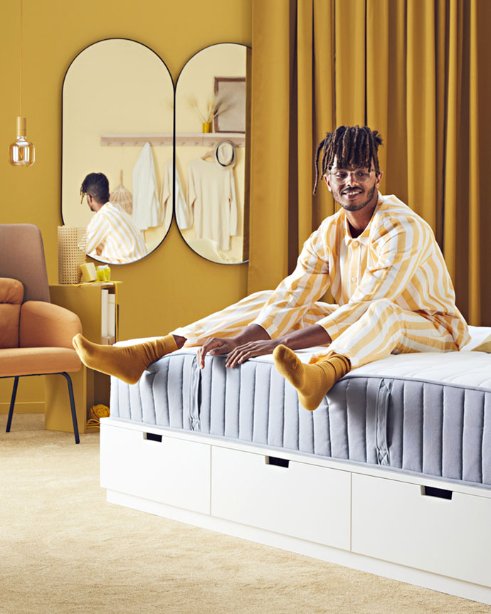 Hombre sonriendo sentado en una cama blanca con cajones, con pijama de rayas naranja/blanco y calcetines amarillos. Habitación amarilla.