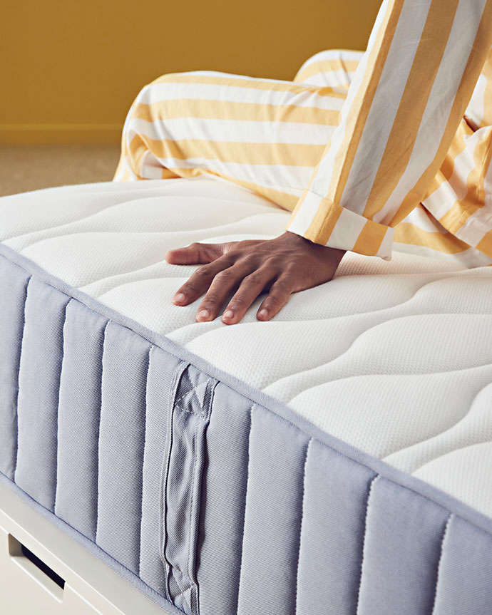 Primer plano de una mano tocando un colchón de muelles ensacados con cubierta blanca y borde gris claro sobre una base de cama con cajones.