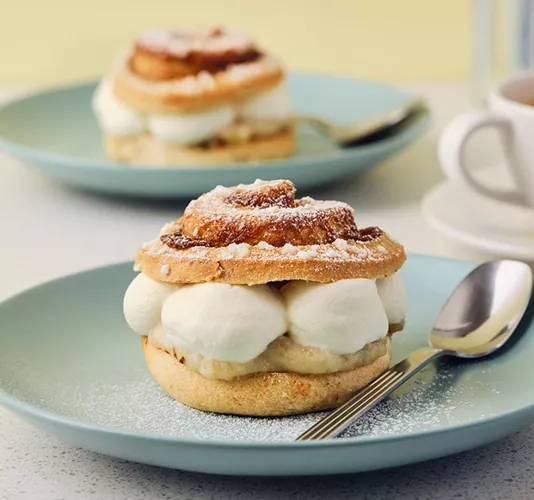 Two cinnamon buns filled with cream on turquoise ceramic plates. The bun in the foreground is dusted with powdered sugar.