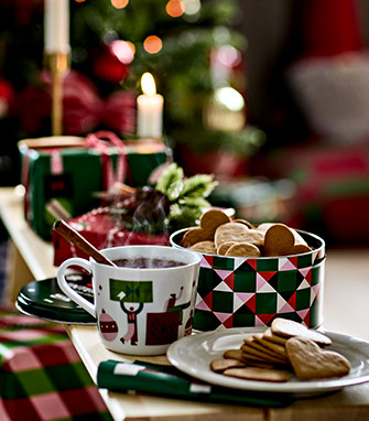 Taza con diseño navideño gráfico, canela y vapor, junto a caja metálica y plato con galletas de jengibre. Árbol de Navidad de fondo.	