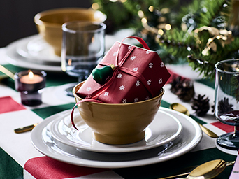 A gift wrapped in red wrapping paper, placed inside a bowl on a Christmas table with white and gold tableware.