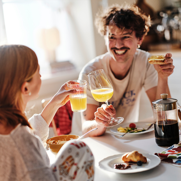 Pareja sonriendo y brindando con zumo de naranja en una mesa de comedor blanca y redonda, con platos, un cruasán y una prensa francesa de café.