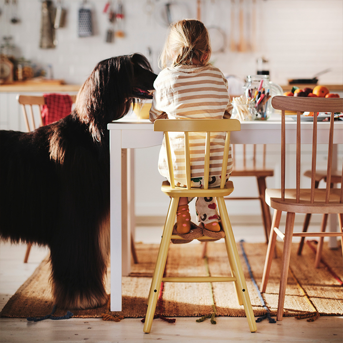 Niña con pijama sentada en una trona infantil de madera de haya teñida de color amarillo en una cocina, junto a una mesa blanca, una silla de madera natural y un perro afgano.